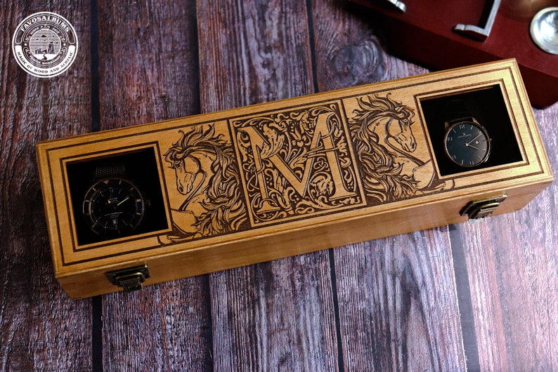 May include: A wooden box with a hinged lid, engraved with a decorative floral design and the letter "M". The box has three compartments, each with a cutout for a watch. Two watches are visible, one with a black face and the other with a silver face.