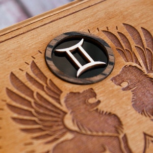 May include: A wooden box with a carved design of two winged lions. The box has a circular inlay with the Gemini zodiac symbol.