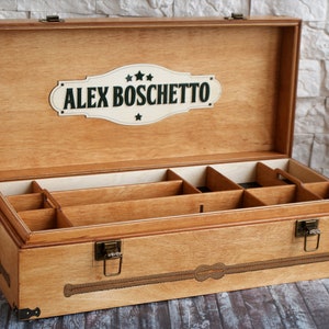 May include: Wooden storage box with multiple compartments, featuring a light brown finish. The box has a hinged lid and metal clasps. The lid displays the name "ALEX BOSCHETTO" on a decorative plaque. The box is designed for organizing items.
