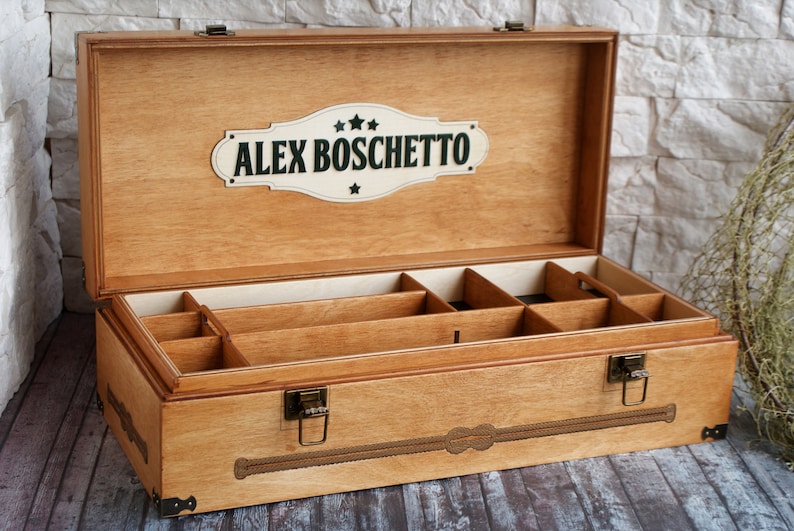 May include: Wooden storage box with multiple compartments, featuring a light brown finish. The box has a hinged lid and metal clasps. The lid displays the name "ALEX BOSCHETTO" on a decorative plaque. The box is designed for organizing items.