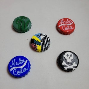 May include: Five bottle caps painted with different designs. One is green with a fern design, one is red with white text that says "Nuka Cola", one is blue with white text that says "Nuka Cola", one is black with a white skull and crossbones, and one is yellow with a multi-colored patchwork design.