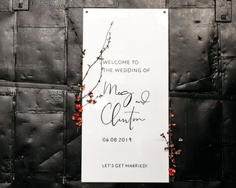 May include: White wedding sign with black text that reads "Welcome to the wedding of Meg and Clinton 06.08.2019 Let's get married!" with a small branch of red flowers on the left side.