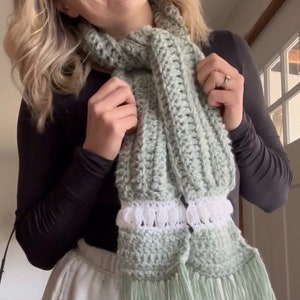 May include: A light green crocheted scarf with white crocheted trim and long fringe. The scarf is worn by a person with blonde hair.