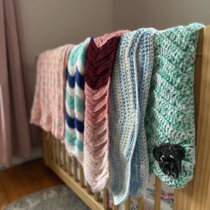 Op de afbeelding: Vijf gehaakte babydekens in verschillende kleuren en patronen hangen aan een wieg. De dekens zijn roze, blauw, groen, wit en bordeauxrood.