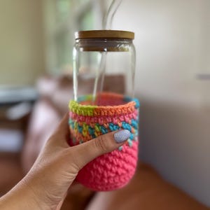 May include: A glass jar with a wooden lid and a crocheted pink, green, blue, and yellow cozy. The jar has two clear drinking straws inside.
