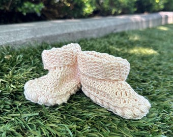 Könnte beinhalten: Ein Paar hellrosa gehäkelte Babyschuhe. Die Schuhe sind aus weichem Garn gefertigt und haben einen umgeschlagenen Bund. Sie liegen auf grünem Gras.
