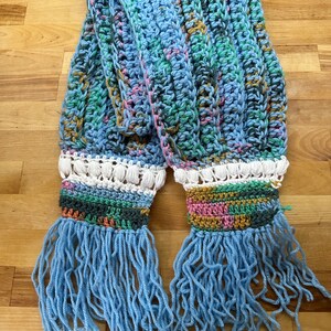 May include: A blue crocheted scarf with colourful stripes and fringe. The scarf has a white crocheted trim with a multi-coloured stripe.