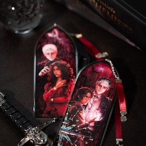 May include: Two gothic-style bookmarks with illustrations of characters in red and black. Each bookmark has a red ribbon attached. A decorative sword and a crystal ball are also visible.