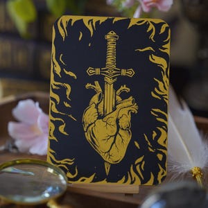 Peut inclure: Un journal noir et or avec une illustration d'épée perçant un cœur. Le design est encadré de motifs en forme de flammes. Le journal est posé sur un présentoir en bois, avec une loupe et des fleurs en arrière-plan. Idéal pour l'écriture.