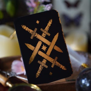 Puede incluir: Una carta de tarot negra con una ilustración dorada de espadas cruzadas y estrellas. La carta es parte de una baraja, probablemente utilizada para la adivinación o prácticas espirituales. El diseño es detallado y visualmente llamativo.
