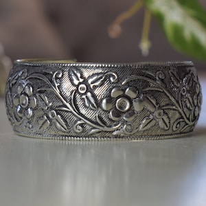 May include: A silver metal bangle bracelet with an intricate floral design. The bracelet is wide and has a rounded shape.