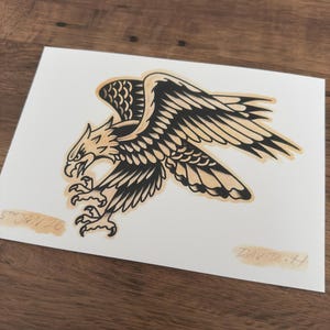 May include: A black and tan eagle illustration on white paper. The eagle is depicted with outstretched wings and sharp talons. The artwork is signed "DAVID-H" and dated "5th JAN 26".