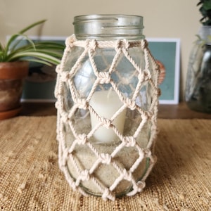 May include: A clear glass jar with a beige macrame net, filled with sand and a white candle. The jar sits on a woven jute mat, creating a coastal or bohemian aesthetic. The jar is a decorative item.
