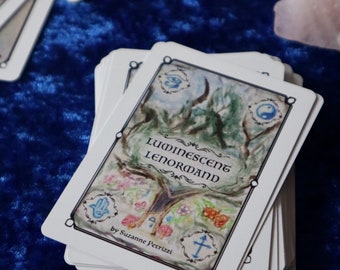 May include: A deck of Luminescent Lenormand cards by Suzanne Petrizze. The cards are white with black borders and feature colorful illustrations. The top card shows a tree with symbols like an eye, a yin yang, a hand, a house, and a cross.
