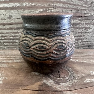 May include: A small, round, dark brown and black ceramic pot with a wide, flared rim. The exterior features a textured, repeating wave-like pattern carved into the clay, with dark glaze accents highlighting the recessed areas of the design.