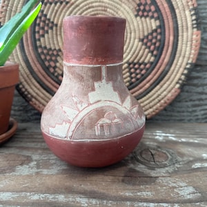 May include: A small, handcrafted terracotta vase with a rounded base and a cylindrical neck. The vase features a carved design of a sun and geometric shapes in white against a reddish-brown background. The vase is displayed on a wooden surface.