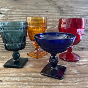 May include: Four colorful vintage glassware pieces. The glasses are in shades of teal, amber, red, and cobalt blue. Each glass has a unique design, including diamond and textured patterns. They sit on square bases, showcasing a variety of stem styles.