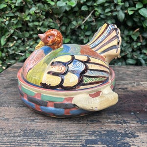 May include: A hand-painted ceramic turkey-shaped lidded casserole dish. The base features a woven basket pattern in blue, red, and green. The lid is shaped like a turkey with yellow, blue, and brown feathers and a prominent handle.