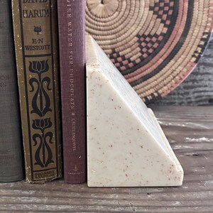 May include: A triangular bookend made of off-white stone with small brown speckles throughout. The bookend is positioned next to two hardcover books, one titled David Harum by E.N. Westcott and the other Like Water for Chocolate by Laura Esquivel.