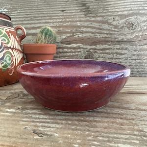 May include: A handmade, glazed ceramic bowl with a rich burgundy color and a glossy finish. The bowl has a wide, shallow shape, perfect for serving or display. The bowl is set on a rustic wooden surface.
