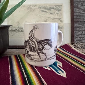 May include: A cream-colored ceramic mug featuring a black ink illustration of a person wearing a cowboy hat riding a horse. The drawing includes a saddle and reins, with the signature Southgate visible at the bottom of the graphic design.