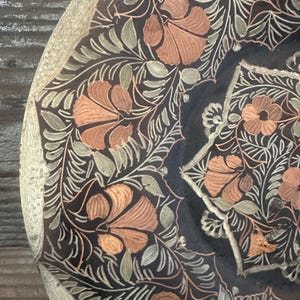 May include: Close-up of a decorative plate with an intricate floral design. The plate features a dark background with copper-colored flowers and green leaves. The rim of the plate has a light beige color.