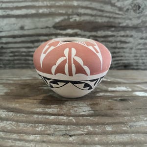 May include: A small, round, terracotta-colored pottery vessel with a narrow opening. The vessel features white and black geometric designs, including a band of triangles and stylized shapes. The background is a weathered wooden surface.