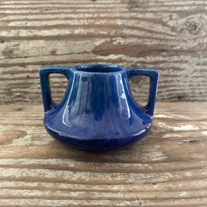 May include: A small, blue ceramic vase with two handles. The vase has a glossy finish and a rounded base. The color is a deep, rich blue with lighter blue streaks. The vase is sitting on a wooden surface.