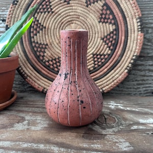 May include: A terracotta-colored ceramic vase with vertical grooves and black speckles. The vase has a rounded base and a narrow neck. A woven basket and a potted plant are in the background, set on a weathered wooden surface.