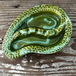 May include: Green ceramic ashtray with a textured surface and a unique, wavy design. The ashtray has two compartments for cigarettes.
