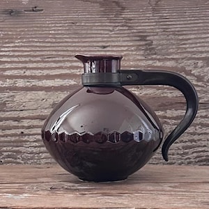 May include: A maroon ceramic coffee pot with a black handle and lid. The pot has a rounded body with a decorative honeycomb pattern around the base. The handle is curved and attached to the top of the pot.