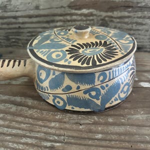 May include: A small, round ceramic pot with a lid. The pot is painted with blue and black floral patterns on a cream background. It has a handle on one side and a small knob on the lid. The pot is likely used for cooking or serving.