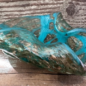 May include: A handcrafted glass bowl with a flowing, organic shape. The bowl features a blend of turquoise and gold colors, creating a shimmering effect. The glass appears translucent, revealing the intricate patterns within.