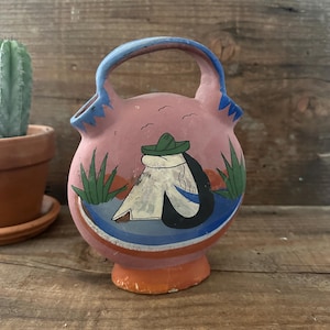 May include: A pink ceramic pitcher with a handle, featuring a hand-painted scene of a person in a sombrero. The design includes blue, green, and orange accents. The pitcher has a rounded body and a small, orange base.