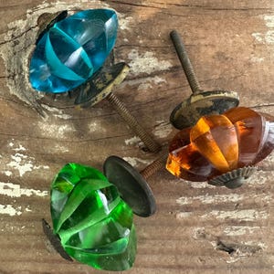 May include: Three decorative glass cabinet knobs in teal, amber, and green. Each knob has a faceted design and is attached to a metal screw. The knobs are set against a weathered wooden surface.