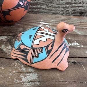 May include: A terracotta-colored clay turtle figurine featuring a hand-painted shell with geometric patterns in turquoise, black, and white. The turtle has a smooth, rounded body, a small head with black painted eyes, and four short, stubby legs.