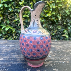 May include: A ceramic pitcher with a tan base and a rounded body featuring a red section decorated with blue floral patterns and wavy lines. The neck displays black painted grass motifs, while the upper shoulder has a band of circular floral designs.