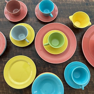 May include: A collection of colorful ceramic dishware, including plates, saucers, cups, and a bowl. The set features various colors, including red, yellow, green, blue, and gray. The arrangement is on a dark wooden surface.