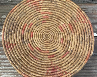 May include: Round woven basket with a spiral design. The basket is made of natural-colored fibers with accents of red and gray. The basket's circular shape and intricate weave create a visually appealing texture. The basket is likely handmade.