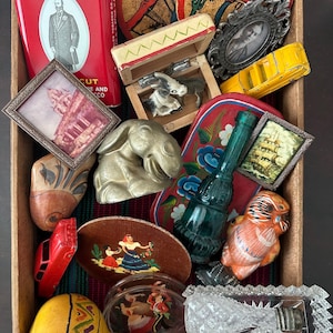 May include: A wooden box filled with various vintage items. Includes a red Prince Albert tobacco tin, framed photos, decorative coasters, a small wooden box with donkey figurines, a gold elephant figurine, and a glass bottle.