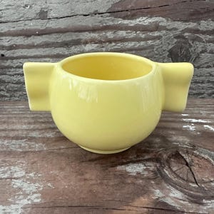 May include: A small, round, pale yellow ceramic bowl featuring two wide, flat, rectangular handles extending horizontally from opposite sides. The bowl has a smooth, glossy finish and a simple, open-top design with a consistent, uniform color.