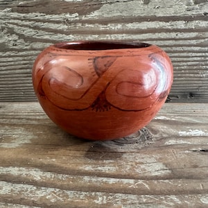May include: A round, terracotta-colored pottery bowl with a dark brown interior. The exterior features a repeating, stylized design in dark brown. The bowl sits on a weathered wooden surface, showcasing its rustic charm.