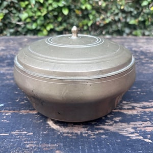 May include: A round, metallic brass-toned container with a matching lid. The lid features a small, centered knob handle and concentric circular etched lines. The base of the container has a smooth, curved profile with a slightly wider top rim.