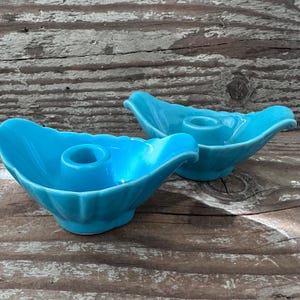 May include: Two turquoise ceramic candle holders. Each holder has a petal-like shape with a central circular opening for a candle. The holders are placed on a weathered wooden surface.