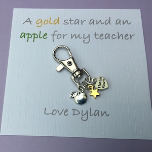 Op de afbeelding: Een witte kaart met de tekst "A gold star and an apple for my teacher" en "Love Dylan". Een zilveren sleutelhanger met een zilveren appelbedel, een gouden sterbedel en een zilveren hartbedel met de tekst "Thank you".