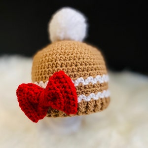 May include: A brown crocheted hat with a white pom-pom on top and a red crocheted bow on the side.