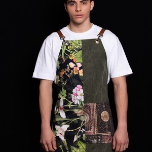 May include: An olive green apron with a floral and patterned design. The apron features a patchwork design with a section of dark floral print and a section of patterned fabric. The apron has brown leather straps and metal grommets.