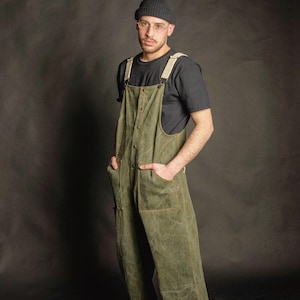Pode incluir: Jardineiras de lona verde-oliva com carcela de botões frontal, dois bolsos grandes e alças de cor clara. A peça é usada sobre uma t-shirt preta lisa de manga curta, combinada com um gorro cinzento-escuro e sapatilhas brancas.