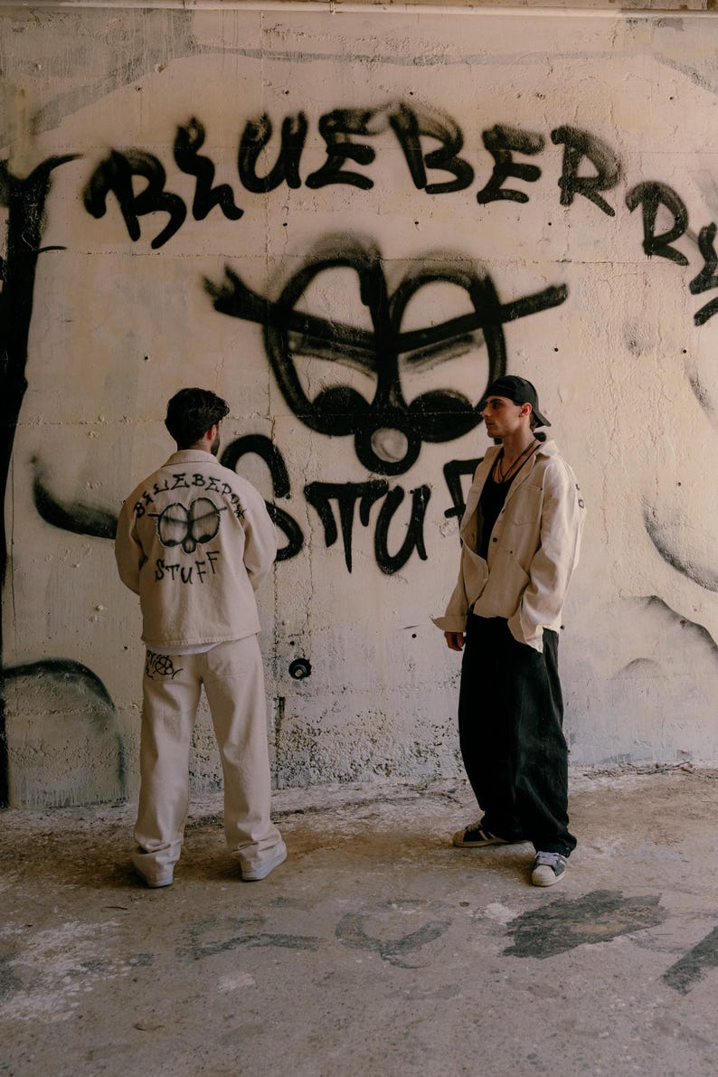 May include: Two individuals wearing beige button-down shirts and matching beige trousers, with one shirt featuring a graphic print on the back that reads "BLUEBERRY STUFF" above a stylised circular icon. The other individual wears black trousers.