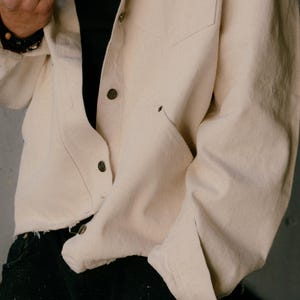 May include: A long-sleeved, off-white button-down shirt made of a heavy fabric, featuring a chest pocket, a pointed collar, and metallic buttons. The shirt has a raw, unfinished hemline at the bottom and is worn over a plain black undershirt.